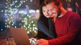 Ein Mann sieht lachend auf den Bildschirm eines Laptops. Eine Frau ist angelehnt an seiner Schulter.