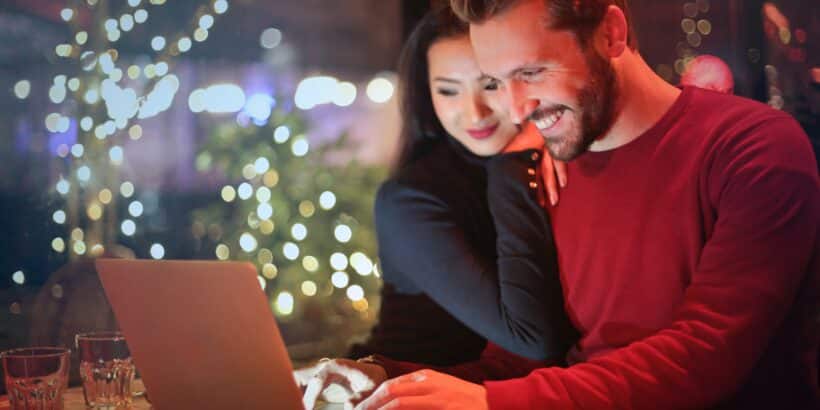 Ein Mann sieht lachend auf den Bildschirm eines Laptops. Eine Frau ist angelehnt an seiner Schulter.